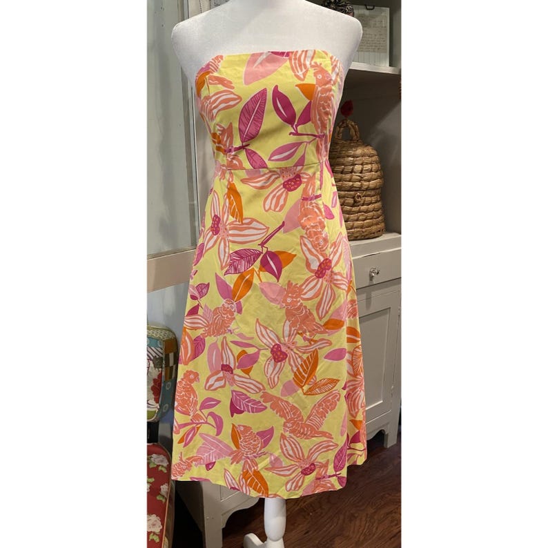 May include: A strapless, knee-length dress with a vibrant floral pattern. The dress features a yellow background with large pink, orange, and white flowers and leaves. The dress is displayed on a mannequin.