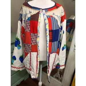 May include: A long-sleeved open-front jacket featuring a patchwork design with various red, blue, and white fabric patterns. The garment includes white panels with blue strawberry prints, two front pockets, and a decorative scalloped trim.