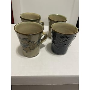 Puede incluir: Un juego de cuatro tazas de cerámica con asa. Dos tazas son de color beige claro con una ilustración de un pez marrón, una es de color beige claro sólido y otra es de color gris carbón oscuro con un patrón floral en relieve en el exterior.
