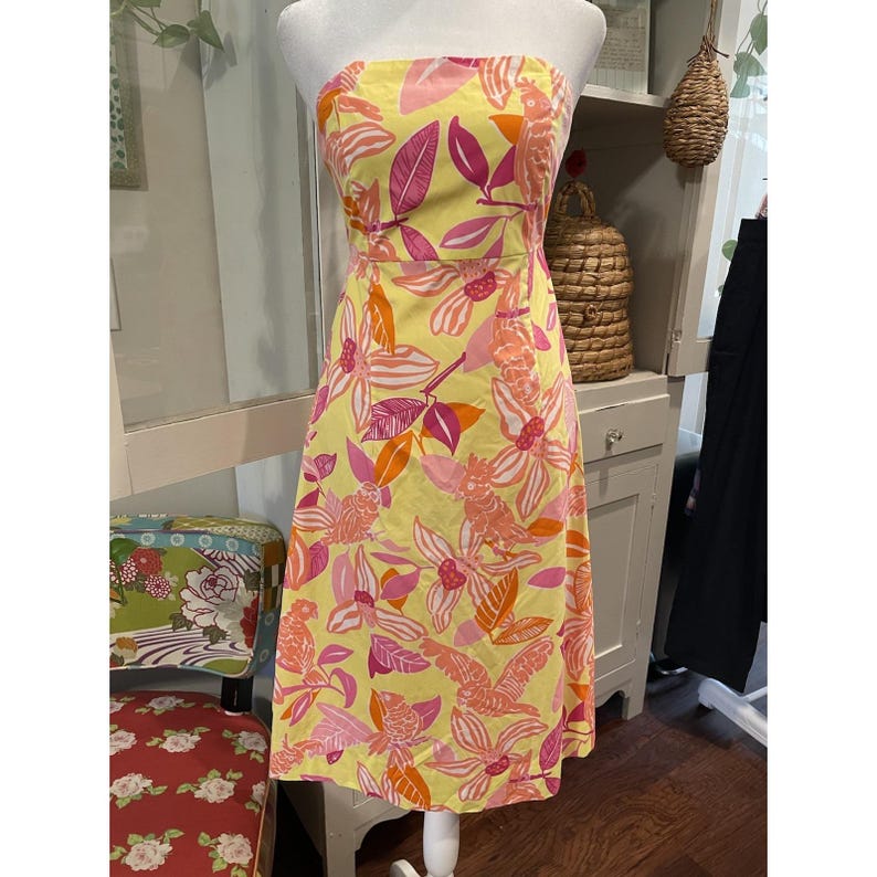 May include: A strapless, knee-length dress in a vibrant yellow hue, adorned with a floral pattern in shades of pink and orange. The dress is displayed on a mannequin, showcasing its design and fit. The floral print includes stylized birds and leaves.