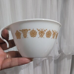 May include: White ceramic cup with a handle and a decorative band of gold floral patterns. The cup has a wide opening and a curved handle. The design is a vintage style, suitable for serving beverages or as a decorative piece.