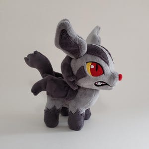 Puede incluir: Un peluche con pelaje gris claro y oscuro, orejas grandes y puntiagudas y una pequeña nariz roja. Presenta ojos amarillos y rojos, una boca pequeña con dientes blancos visibles y una cola corta. El juguete de tela se sostiene sobre cuatro patas.