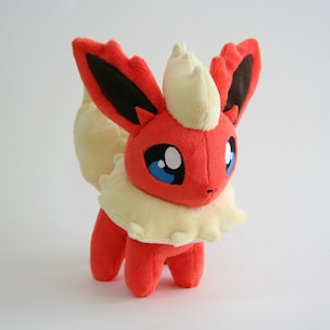Puede incluir: Un juguete de peluche de un Pokémon parecido a un zorro rojo y amarillo con ojos azules y un cuello de color crema alrededor del cuello.