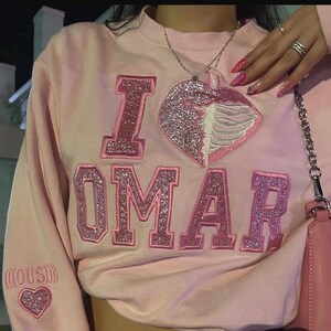 K&ouml;nnte beinhalten: Hellrosa Sweatshirt mit den Worten "I OMAR" in glitzernden rosa Buchstaben. Ein herzf&ouml;rmiger Aufn&auml;her mit einem Rippenmuster befindet sich auf der Vorderseite. Ein kleines Herz ist auf dem &Auml;rmel gestickt.