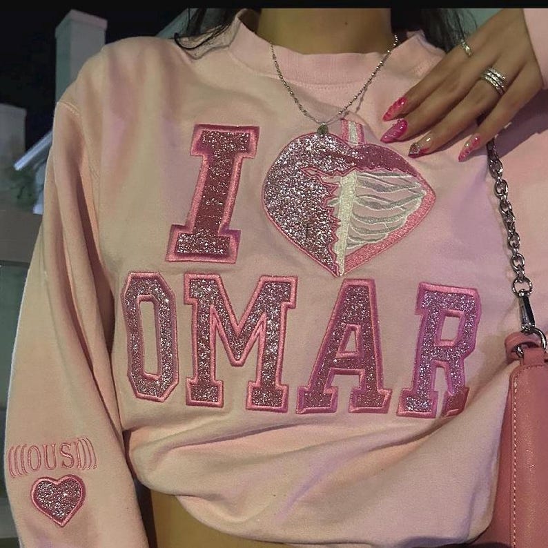 K&ouml;nnte beinhalten: Hellrosa Sweatshirt mit den Worten "I OMAR" in glitzernden rosa Buchstaben. Ein herzf&ouml;rmiger Aufn&auml;her mit einem Rippenmuster befindet sich auf der Vorderseite. Ein kleines Herz ist auf dem &Auml;rmel gestickt.