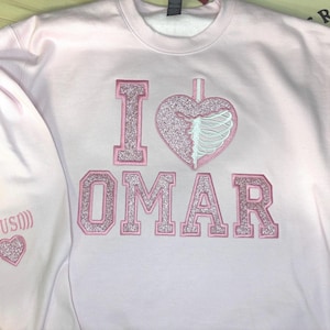 K&ouml;nnte beinhalten: Ein hellrosa Sweatshirt mit den gestickten Worten "I OMAR" in glitzerndem rosa Schriftzug. Das "I" ist ein Blockbuchstabe, und das Herz hat ein Rippendesign. Der linke &Auml;rmel hat ein Herz und das Wort "(OUSI)" gestickt.