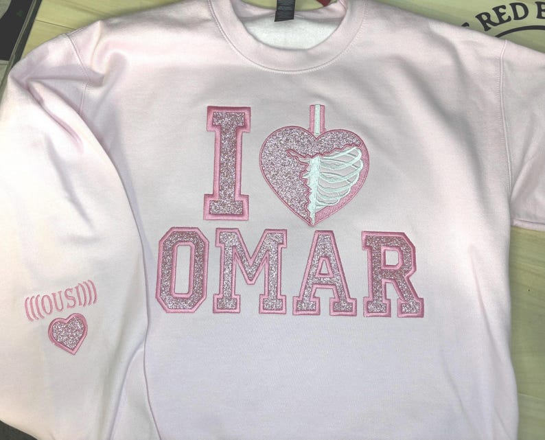 K&ouml;nnte beinhalten: Ein hellrosa Sweatshirt mit den gestickten Worten "I OMAR" in glitzerndem rosa Schriftzug. Das "I" ist ein Blockbuchstabe, und das Herz hat ein Rippendesign. Der linke &Auml;rmel hat ein Herz und das Wort "(OUSI)" gestickt.