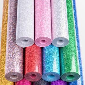 K&ouml;nnte beinhalten: Eine Sammlung gerollter Glitzer-Vinyls in verschiedenen Farben, darunter Gold, Rosa, Rot, Blau und Gr&uuml;n. Die Rollen sind eng gewickelt und zeigen die glitzernde Textur des Materials. Das Bild ist gut beleuchtet und hebt die reflektierenden Eigenschaften des Glitzers hervor.