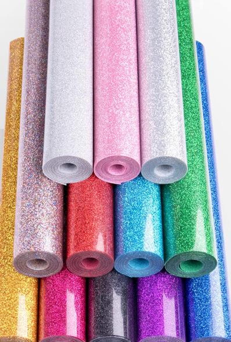 K&ouml;nnte beinhalten: Eine Sammlung gerollter Glitzer-Vinyls in verschiedenen Farben, darunter Gold, Rosa, Rot, Blau und Gr&uuml;n. Die Rollen sind eng gewickelt und zeigen die glitzernde Textur des Materials. Das Bild ist gut beleuchtet und hebt die reflektierenden Eigenschaften des Glitzers hervor.