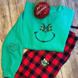 May include: Green sweatshirt with a Grinch face and a red heart on the sleeve. The sweatshirt is paired with red and black plaid pajama pants.