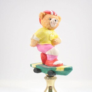 May include: A ceramic lamp finial featuring a teddy bear wearing a red and white helmet, a yellow shirt, pink pants, and red shoes, riding a green and yellow skateboard.