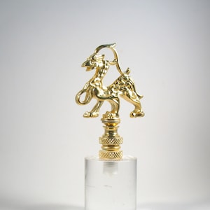 May include: A gold-colored lamp finial featuring a stylized griffin design. The griffin has a bird-like head and the body of a lion. The finial is mounted on a clear, cylindrical base. The finial is made of metal.