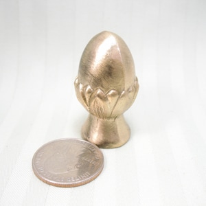 May include: A gold-colored metal acorn finial with a detailed design. The finial is about the size of a quarter.