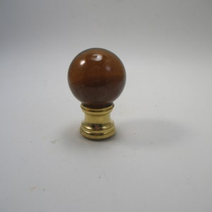 May include: A brown glass lamp finial with a gold metal base. The finial is round and has a smooth surface.