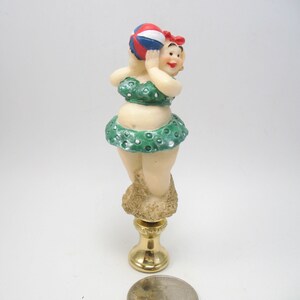 May include: A green and white polka dot bikini-clad figurine holding a red, white, and blue beach ball. The figurine is standing on a sand-colored base with a gold lamp finial.