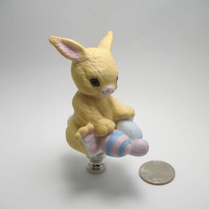 May include: A yellow ceramic bunny lamp finial with pink ears and blue and pink striped eggs. The finial is sitting on a silver base.