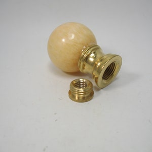May include: A light beige lamp finial with a spherical top and a brass base. The finial is separated from a brass threaded nut. The lamp finial is designed to be screwed onto a lamp.