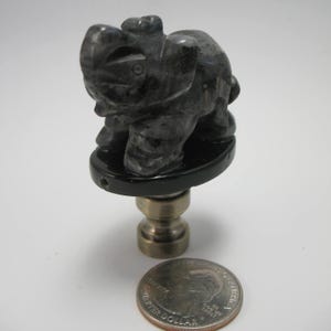 May include: A dark gray carved stone elephant figurine lamp finial. The elephant is standing on a black base, with a brass-colored metal stem. A U.S. quarter coin is next to the finial for size comparison.