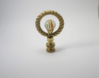 May include: A brass lamp finial featuring a decorative rope-like ring surrounding a clear marble with orange and white swirls. The finial has a detailed, textured base and is designed to top a lamp.