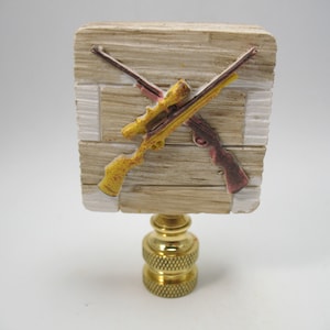 May include: A lamp finial with a wooden base and two crossed rifles. The rifles are painted in yellow and red. The base is painted to look like weathered wood.