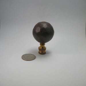 May include: A dark brown, spherical lamp finial with a textured surface, mounted on a brass base. A coin is next to the finial for size comparison. The finial is a decorative element for lamps.