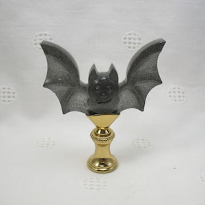 May include: A decorative bat figurine with outstretched wings, in shades of gray and black, atop a gold-colored base. The bat has a smiling face and a speckled texture. A unique Halloween or gothic-themed accent piece.