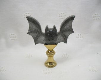 Könnte beinhalten: Dekorative Fledermausfigur in Grau und Schwarz mit ausgebreiteten Flügeln, auf einem goldfarbenen Sockel. Die Fledermaus hat ein lächelndes Gesicht und eine gesprenkelte Textur. Ein einzigartiges Halloween- oder Gothic-Themen-Accessoire.