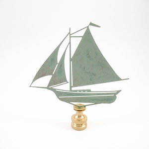 May include: A decorative sailboat lamp finial. The sailboat is cut from a weathered green metal and is mounted on a brass base. The sailboat has two sails and a mast. The finial is designed to be used on a lamp.