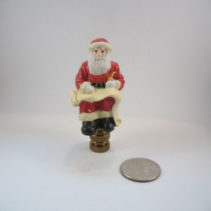 May include: A red and white Santa Claus figurine sitting on a gold lamp finial. He is holding a scroll and a pipe. The figurine is about 2 inches tall.