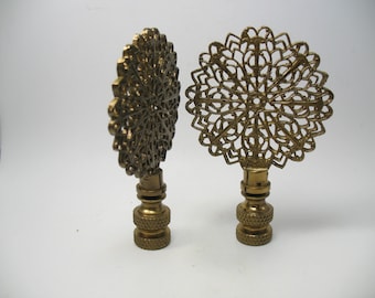 May include: Two brass-colored lamp finials. Each finial features a decorative, openwork design in a circular shape. The base of each finial is a textured, cylindrical shape. The finials are set against a white background.