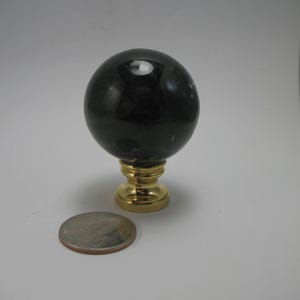 May include: A dark, spherical lamp finial with a gold-colored base. A US quarter is next to the finial for size comparison. The finial has a polished, reflective surface.
