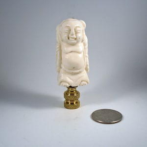 May include: Ivory-colored lamp finial featuring a smiling Buddha figure. The Buddha is seated with hands resting on the lap. The finial is mounted on a brass base. A quarter coin is next to the finial for size comparison.