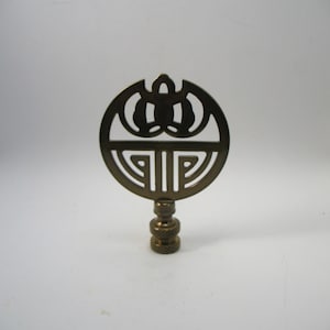 May include: A gold-toned metal lamp finial with a circular design featuring a stylised geometric pattern. The finial has a threaded base for attaching to a lamp.