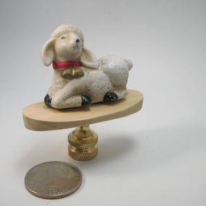 May include: A lamp finial featuring a ceramic lamb figurine with a red collar and gold bells. The lamb is resting on a wooden base, supported by a brass fitting. A quarter is next to the finial for size comparison.