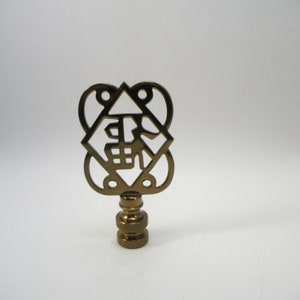May include: Gold metal lamp finial with a decorative cutout design featuring a Chinese character. The finial is approximately 2 inches tall.