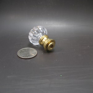 May include: A clear, ribbed glass knob with a brass base. The knob is next to a quarter coin for size comparison. The knob is likely for a drawer, cabinet, or furniture piece.