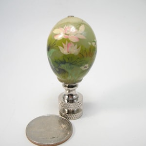 May include: An oval-shaped lamp finial with a painted floral design in shades of pink and green. The finial is mounted on a silver-colored, textured base. A U.S. quarter is next to the finial for size comparison.