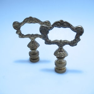 May include: Two ornate brass lamp finials with a decorative openwork design. The finials have a detailed, scroll-like pattern around the open center and a textured base. The overall design is vintage and elegant.