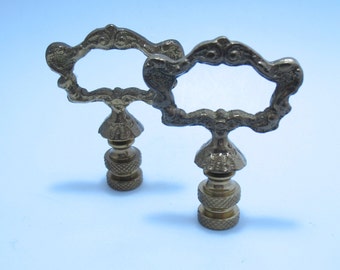 May include: Two ornate brass lamp finials with a decorative openwork design. The finials have a detailed, scroll-like pattern around the open center and a textured base. The overall design is vintage and elegant.