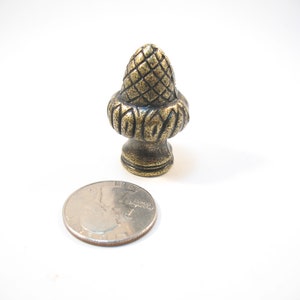 May include: A gold-toned metal finial with a detailed acorn design. The finial is about the size of a US quarter.
