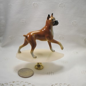 May include: A porcelain figurine of a brown and white boxer dog standing on a white alabaster base with a gold metal finial.