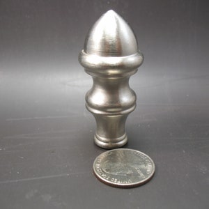 May include: A silver metal finial with a pointed top and a decorative design. The finial is about the size of a US quarter.