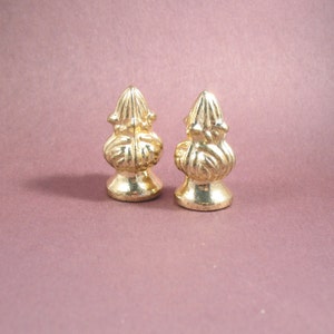 May include: Two gold-colored metal finials with an ornate design. The finials are on a pink background.
