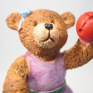 May include: A brown teddy bear wearing a purple dress and a blue bow is holding a red ball.