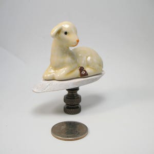 May include: A ceramic lamp finial featuring a white lamb with brown accents. The lamb is lying down and has a black eye. The finial is attached to a brass lamp base.