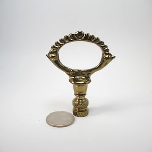 May include: A brass lamp finial with an ornate oval design and a textured base. The finial is approximately 3 inches tall, with a decorative oval top and a threaded base for attachment to a lamp.
