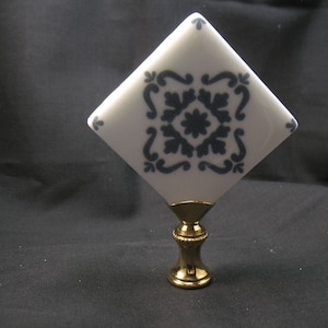 May include: A white and navy blue ceramic lamp finial with a diamond shape. The finial features a navy blue floral pattern and a gold-colored base. The finial is a decorative element for lamps.