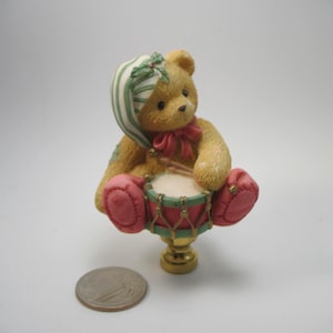 May include: A ceramic Christmas light pull featuring a teddy bear wearing a green and white striped hat and playing a drum. The bear is sitting on a gold base.