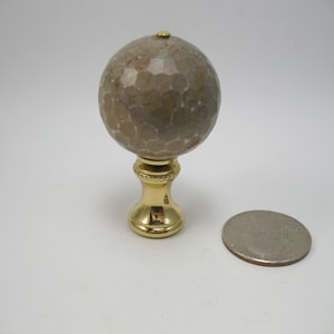 May include: A gold lamp finial with a round, beige, textured top. The finial is about the size of a US quarter.