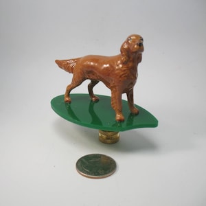 May include: A brown dog ornament standing on a green, leaf-shaped base. The figurine is detailed and mounted on a brass-coloured stem. A coin is placed below the ornament for size comparison.